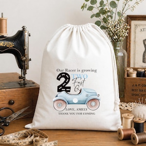 Puede incluir: Una bolsa de tela blanca con cordón que presenta un gráfico impreso de un coche de carreras vintage de color azul claro. El texto dice: Our Racer is growing 2 Fast, LOVE, AMELY, THANK YOU FOR COMING. La bolsa tiene un cierre fruncido.