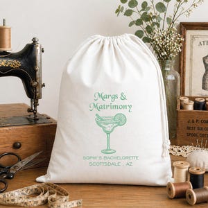 May include: A white drawstring bag with the text "Margs & Matrimony" and a margarita graphic in light green. The bag also includes the text "SOPHI'S BACHELORETTE SCOTTSDALE, AZ". The bag is on a wooden surface with sewing supplies.