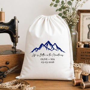 Könnte beinhalten: Ein weißer Kordelzugbeutel mit einem blauen Bergmotiv und dem Text "Life is Better in the Mountains." Der Beutel enthält auch die Namen "OLIVE + NIA" und das Datum "03-03-2026."