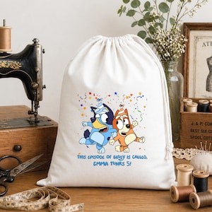 Op de afbeelding: Een witte tas met trekkoord voorzien van een kleurrijke afbeelding van twee tekenfilmhonden met feesthoedjes, omringd door confetti. Onder de figuren staat de tekst: THIS EPISODE OF BLUEY IS CALLED, EMMA TUNRS 5! De tas heeft een gesloten bovenkant.