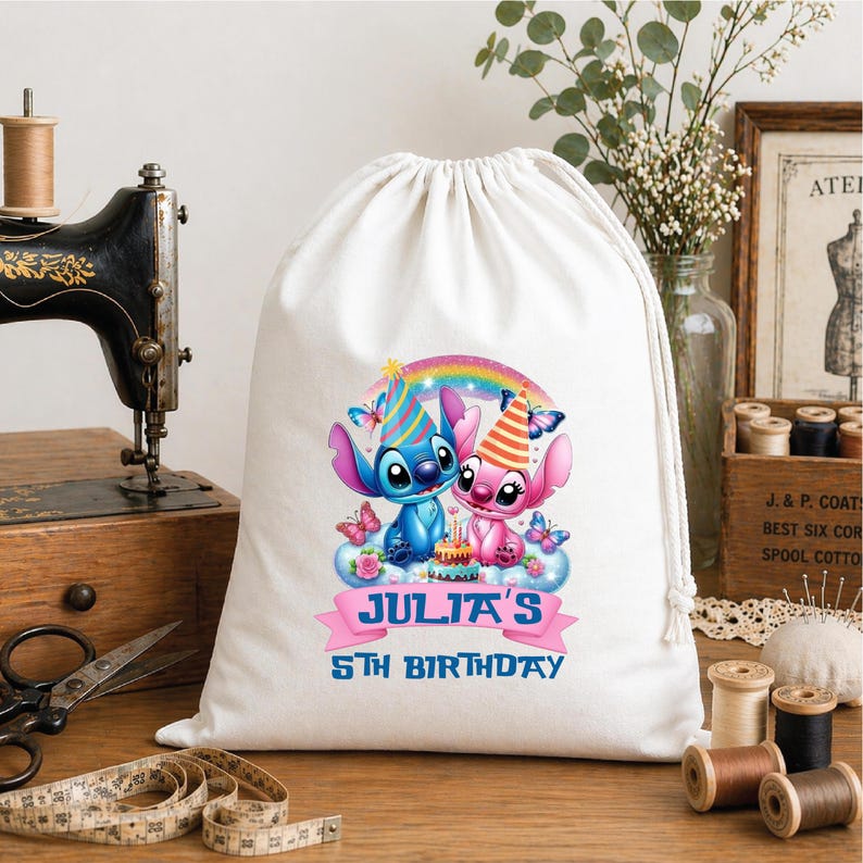 May include: A white drawstring bag featuring a colorful graphic of two cartoon characters, a blue one and a pink one, with party hats, a rainbow, and butterflies. The text "JULIA'S 5TH BIRTHDAY" is displayed on a pink banner.