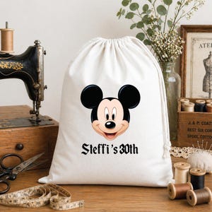 Peut inclure: Un sac en coton blanc avec cordon de serrage, orné d'un visage souriant de Mickey Mouse, avec des oreilles noires et une bouche rouge. Le texte "Steffi's 30th" est imprimé en dessous.