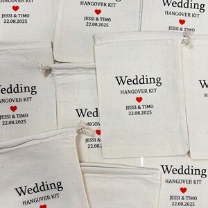 May include: Several off-white fabric bags with drawstrings are displayed. Each bag has the words "Wedding HANGOVER KIT" printed in black, with a red heart graphic. The names "JESSI & TIMO" and the date "22.08.2025" are also printed on each bag.