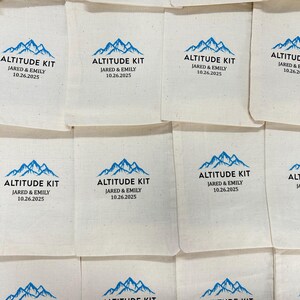 May include: Off-white fabric pouches with a blue mountain graphic and the text "ALTITUDE KIT JARED & EMILY 10.26.2025". The pouches are arranged in a grid pattern. The design is simple and clean.