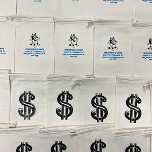 May include: Off-white canvas bags with black printed dollar signs and text. Some bags feature a cartoon character and the text "THIS EPISODE IS CALLED EMMY'S TURNING 4!" in blue. The bags are likely for party favors.