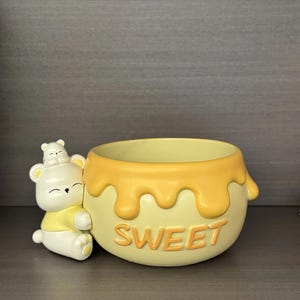 May include: A yellow ceramic bowl with a honey design and the word "SWEET" in orange. A white bear figurine with a smaller bear on its head is attached to the side. The bowl is suitable for small plants.