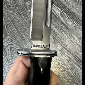 Puede incluir: Primer plano de un cuchillo plateado y negro con la palabra "SCREAM" grabada en la hoja. El cuchillo tiene una hoja brillante y reflectante y un mango negro. El cuchillo está sostenido por una persona.