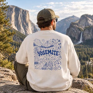 Könnte beinhalten: Ein cremefarbenes Sweatshirt mit Rundhalsausschnitt und einem blauen Grafikdruck auf der Rückseite. Das Design zeigt den Schriftzug YOSEMITE, Berglandschaften, einen Bären, einen Hirsch, ein Zelt, Wanderschuhe, einen Rucksack, ein Fernglas und eine Tasse.