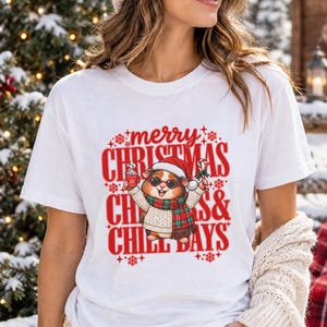 Puede incluir: Una camiseta blanca de manga corta con un gráfico de una cobaya con gorro de Papá Noel, gafas de sol y bufanda de cuadros. La cobaya sostiene una taza y un bastón de caramelo. Texto rojo "merry CHRISTMAS CHILL DAYS" con copos de nieve.