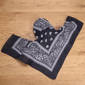 May include: A black bandana featuring a white paisley pattern with a decorative border. The fabric is folded into a triangular shape, displaying the intricate floral and teardrop motifs printed in white ink against the solid black background.