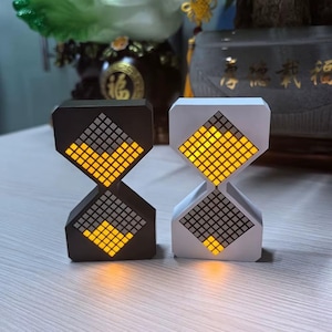 May include: Two hourglass-shaped digital clocks, one black and one white, sit side-by-side. Each clock features a grid of small squares that illuminate in yellow and gray. The clocks have a modern, geometric design.