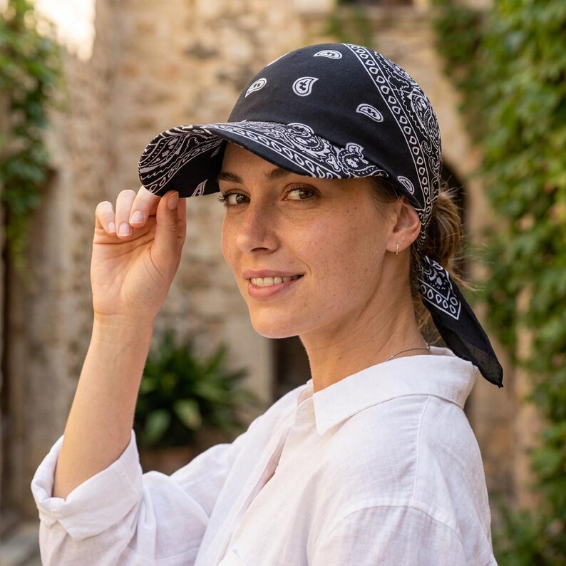 May include: A black baseball cap featuring a white paisley bandana print pattern. The cap has a structured brim and a fabric back that ties at the nape of the neck, extending into two fabric tails that drape down behind the head.