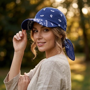 May include: A navy blue baseball cap featuring a white paisley bandana print pattern. The cap has a curved brim and a fabric tie closure at the back, allowing the ends of the bandana-style material to hang down behind the head.