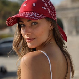 May include: A red baseball cap featuring a white paisley pattern throughout the fabric. The cap has a structured crown and a curved brim, with fabric ties extending from the back, creating a bandana-style appearance in a vibrant red color.