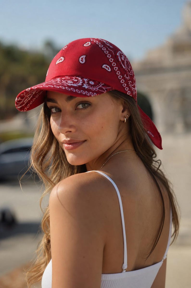 May include: A red baseball cap featuring a white paisley pattern throughout the fabric. The cap has a structured crown and a curved brim, with fabric ties extending from the back, creating a bandana-style appearance in a vibrant red color.