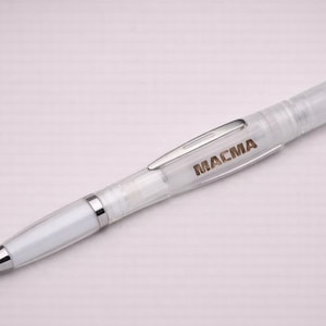 Könnte beinhalten: Ein weißer Stift mit silbernem Clip und Spitze. Auf dem Stift steht das Wort "MACMA". Der Stift liegt auf einem hellrosa Hintergrund.