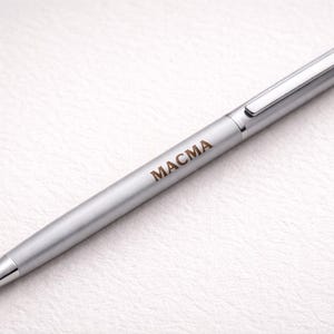 May include: A silver pen with the word "MACMA" printed in brown. The pen has a clip and a pointed tip. The pen is on a white textured surface.