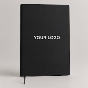 Puede incluir: Un cuaderno rectangular negro con una cubierta mate lisa. El texto "YOUR LOGO" está impreso en blanco, centrado en la portada. Un fino marcapáginas de cinta negra sobresale del borde inferior del cuaderno, que se mantiene en pie.
