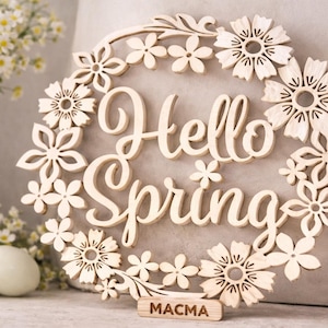 May include: A wooden floral wreath with the words "Hello Spring" cut out in a cursive font. The wreath is decorated with various flower shapes and a small rectangular base with the word "MACMA" engraved on it.