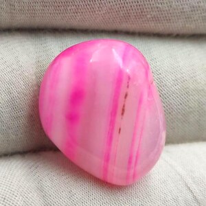 Puede incluir: Una piedra de ágata rosa pulida, de forma ovalada, con bandas de diferentes tonos de rosa y blanco. La piedra tiene una superficie lisa y brillante y algunas pequeñas inclusiones marrones. Objeto decorativo natural.