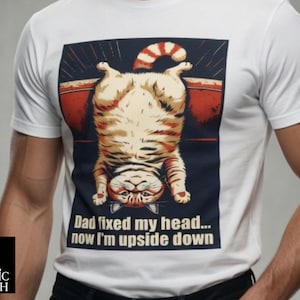 Puede incluir: Camiseta blanca con un diseño de gato de dibujos animados. El gato es naranja y blanco, colgado boca abajo con una cola a rayas. El texto de la camiseta dice "Dad fixed my head... now I'm upside down". La camiseta tiene mangas cortas.