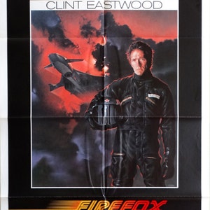 May include: Movie poster for the film "Firefox" starring Clint Eastwood. The poster features a red and black color scheme with a dramatic image of Eastwood in a black flight suit holding a helmet. The text "Clint Eastwood" and "Firefox" are prominently displayed in the poster.