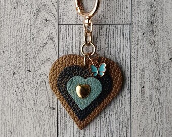 May include: A heart-shaped keychain with a gold clasp and chain. The heart is layered with brown, black, and teal leather, and a small gold heart. A small blue butterfly charm is attached. The keychain is a decorative accessory.