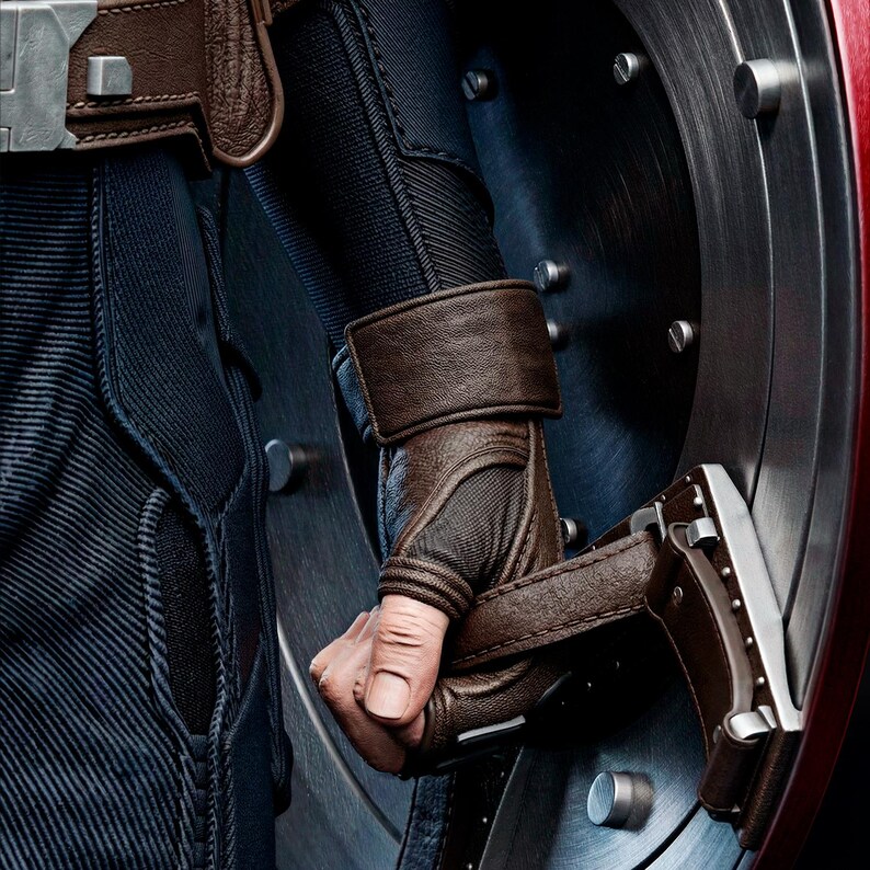 Op de afbeelding: Close-up van een bruine leren handschoen die een zilveren en bruine schildgreep vasthoudt. De handschoen is gedetailleerd met stiksels en een polsband. Het schild is metaalachtig met een rode rand en zilveren accenten. Donkerblauwe stof is zichtbaar.