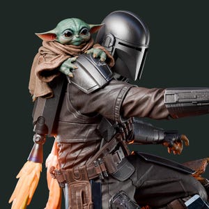 May include: A detailed figurine of The Mandalorian with Grogu. The Mandalorian is wearing a silver helmet and brown armor. Grogu, with green skin and large ears, is perched on his shoulder. A jetpack with orange flames is attached to the Mandalorian's back.