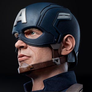 Op de afbeelding: Een gedetailleerd zijaanzicht van een Captain America-helm. De helm is voornamelijk marineblauw met een zilveren "A" op het voorhoofd. Het masker bedekt de ogen en er is een bruine kinriem zichtbaar. De achtergrond is zwart.
