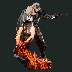 May include: A detailed figurine of a character in a brown and black outfit, with a jetpack and a blaster. The figure is standing on a base with orange and red flames. The character is wearing a helmet and a tan scarf.