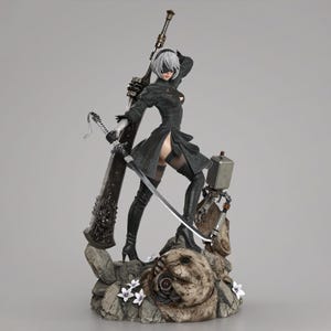 May include: A detailed figurine of a female character from the video game Nier: Automata. She wears a black dress, thigh-high boots, and a blindfold. She holds a large sword and a smaller blade, standing on a rocky base with a small robot.