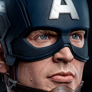 Op de afbeelding: Close-up van een Captain America-buste. De buste heeft een blauwe helm met een zilveren "A"-embleem en een zwart masker. Het personage heeft blauwe ogen en blonde wenkbrauwen. De buste is gedetailleerd met realistische huidtinten en texturen.