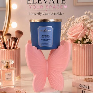 May include: A pink butterfly-shaped candle holder supporting a navy blue candle with gold trim. The candle is labeled "Midnight Blue Citrus." The image includes text that reads "Elevate Your Space" and "Butterfly Candle Holder."