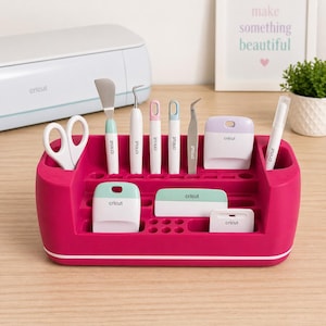 May include: A magenta Cricut tool organiser filled with various crafting tools. Includes scissors, spatulas, weeding tools, a scraper, and a pen. The tools have white, mint green, pink, and lavender accents. A Cricut machine is in the background.