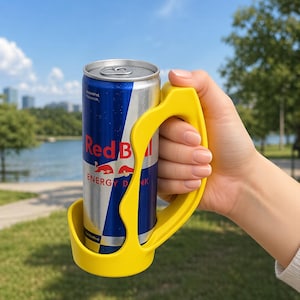 May include: A yellow can holder grips a Red Bull energy drink can. The can is silver and blue with the Red Bull logo. The background shows a lake, trees, and a blue sky.