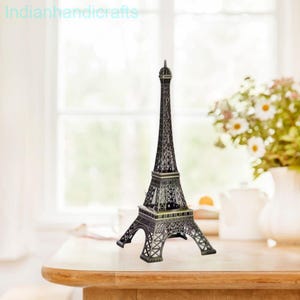 Puede incluir: Una detallada réplica de metal oscuro de la Torre Eiffel. La pieza decorativa se encuentra sobre una mesa con un fondo borroso de una ventana y flores. La torre es un recuerdo y un artículo de decoración popular.