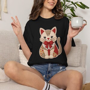 Könnte beinhalten: Ein schwarzes T-Shirt mit einer zentralen Grafik einer stilisierten Katze im Patchwork-Design. Die Katzenillustration enthält verschiedene Muster wie rote Punkte, braunes Vichy-Karo und Blumenprints, abgerundet mit einer roten Schleife.