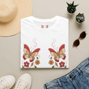 Könnte beinhalten: Ein weißes T-Shirt mit einem Grafikdruck von zwei symmetrischen Schmetterlingen mit rot, gelb und braun gemusterten Flügeln, umgeben von kleinen roten und gelben Blumen, grünen Stielen und gepunkteten Schnörkeln, mit einem kleinen Schmetterlingssymbol am Innenhals.