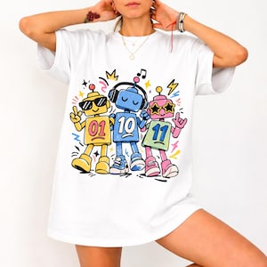 May include: White t-shirt featuring a colorful graphic of three cartoon robots. The robots are yellow, blue, and pink, each with a unique design, sunglasses, and headphones. Each robot has a number: 01, 10, and 11. The design includes musical notes and lightning bolts.