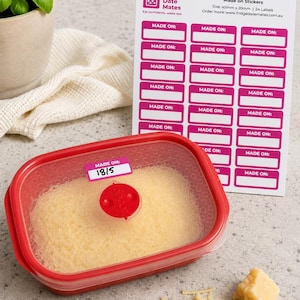 May include: A red food container with grated cheese, labelled with a "Made On" sticker. A sheet of pink and white "Made On" stickers is in the background, with a piece of cheese and a basil plant.