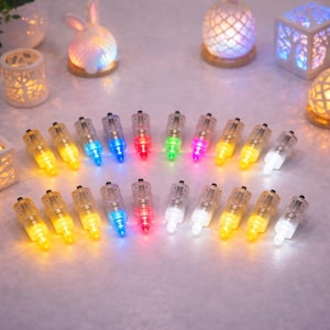 May include: A collection of small, clear LED lights with various colors, including yellow, blue, red, green, and white. The lights are arranged on a light-colored surface, with decorative candle holders and a bunny-shaped light in the background.