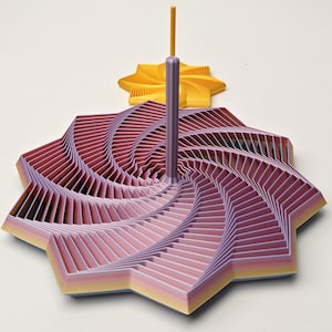 Pode incluir: Uma escultura geométrica em camadas em tons de rosa, roxo e amarelo. Uma haste central roxa eleva-se da estrutura principal, com uma peça menor em forma de estrela amarela no fundo. O design apresenta um padrão em espiral.
