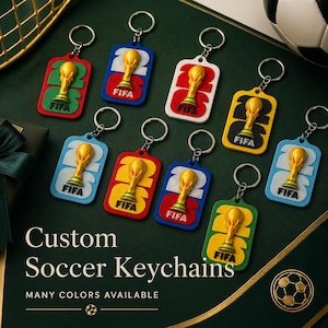 Könnte beinhalten: Eine Sammlung individueller Fußball-Schlüsselanhänger in verschiedenen Farben. Jeder Schlüsselanhänger zeigt ein goldenes Trophäendesign mit dem FIFA-Logo. Die Schlüsselanhänger sind rechteckig mit der Zahl 2.