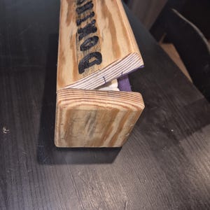 May include: A wooden box with the word "BOXUSES" in black lettering on the top. The box is made of light-coloured wood and has a purple interior. The box is partially open, revealing the interior.