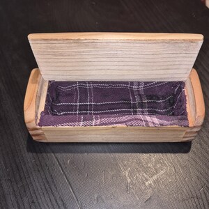 May include: Wooden box with a hinged lid, revealing a purple plaid fabric lining. The box is rectangular with rounded edges and a natural wood finish. The lid is slightly ajar, showcasing the interior.