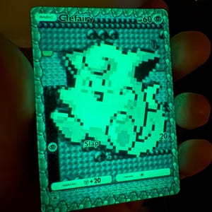 May include: A luminous green Clefairy Pokémon trading card. The card displays a pixelated Clefairy illustration, the word "Slap", and other game text. The card has a textured border and is set against a dark backdrop.
