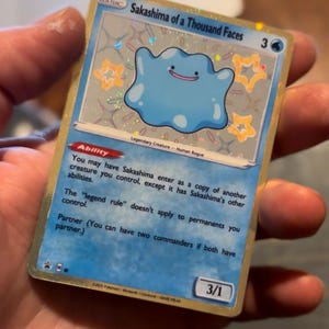 Peut inclure: Une carte à collectionner représentant le personnage Sakashima of a Thousand Faces. La carte a un fond bleu avec un personnage en forme de blob bleu clair et des accents d'étoiles. Le texte comprend "Ability" et "Partner".