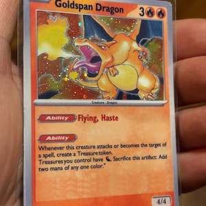 Puede incluir: Carta coleccionable Goldspan Dragon en funda protectora. La carta presenta un dragón naranja con fondo de fuego. Texto: "Goldspan Dragon", "Flying, Haste" y "4/4". La carta es de la serie Basic.