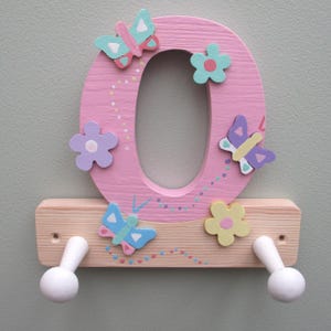 May include: A pink wooden letter "O" decorated with colourful butterflies and flowers. The letter has a wooden shelf with two white knobs below. The design is painted with pastel colours and is mounted on a light green wall.